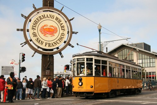Image shows the historic F Line streetcar arriving at Fisherman’s Wharf, San Francisco. Visitors line up to board.