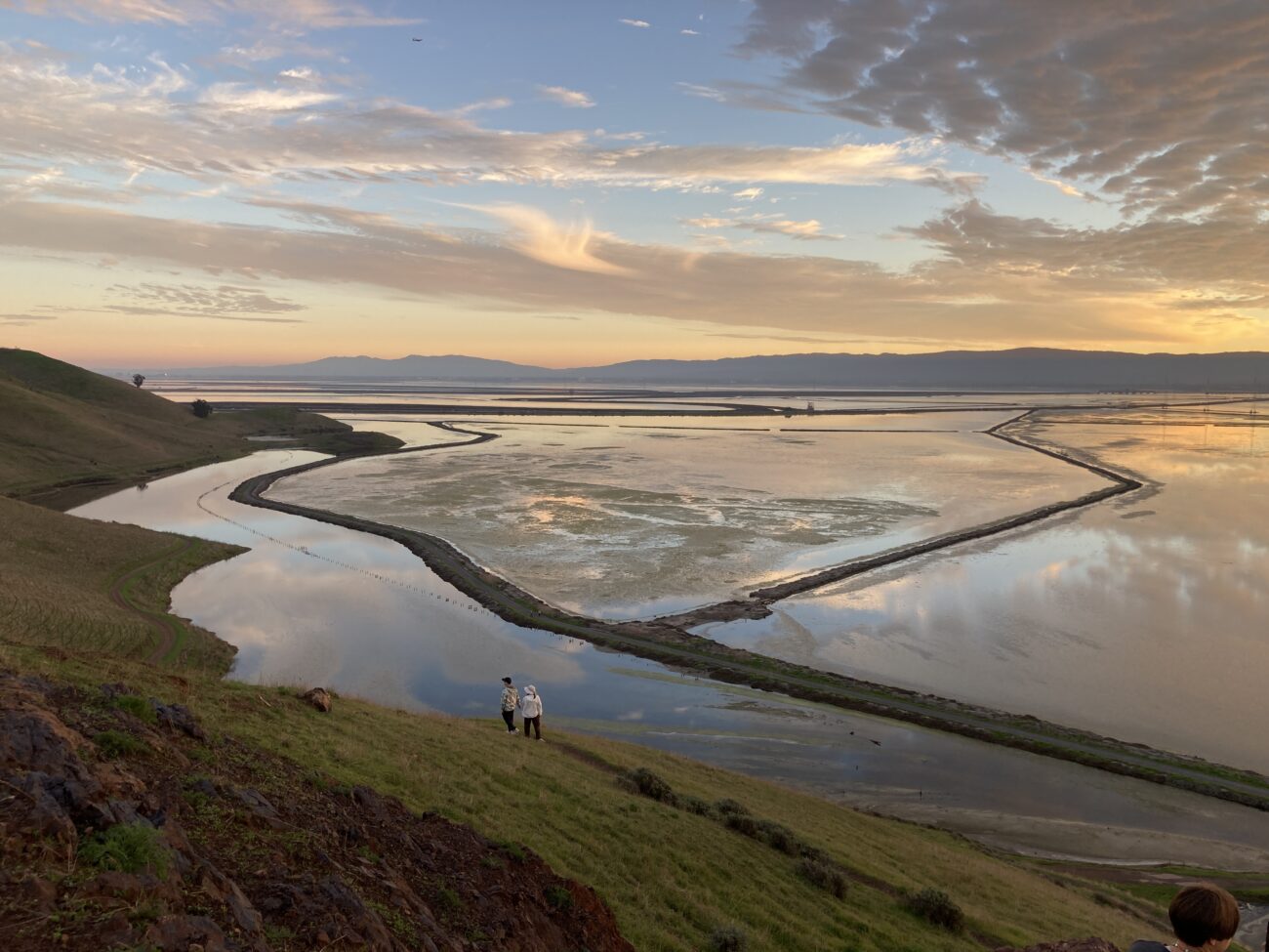 two people walking along hillside trail in front of large wetalnad area at sunset